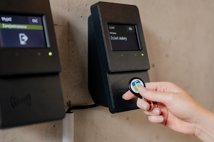 Osoba przykłada chip RFID do cyfrowego terminala rejestracji czasu pracy. Na polskojęzycznych wyświetlaczach widoczne są między innymi komunikaty „Dzień dobry”, „Wyjdź” i „Zarejestrowano”.